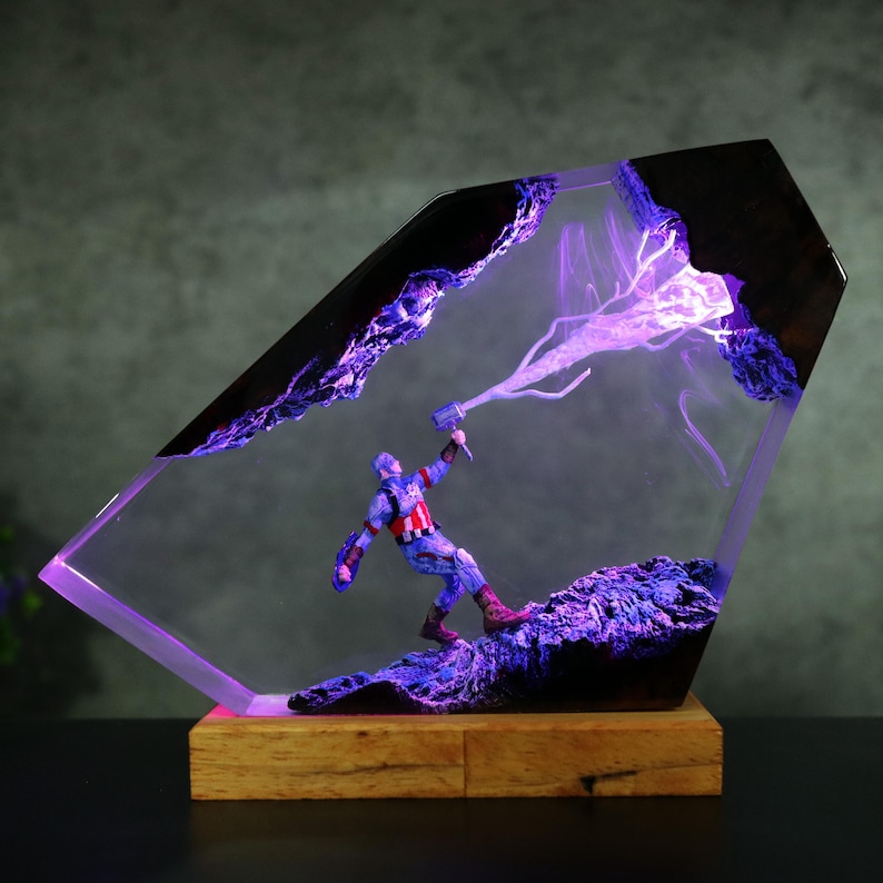 K&ouml;nnte beinhalten: Dekorative Resin-Skulptur mit Captain America und seinem Hammer, eingebettet in einen geometrischen Rahmen. Die Skulptur hat einen Holzsockel und wird mit violetter Beleuchtung beleuchtet, was einen dramatischen Effekt erzeugt.