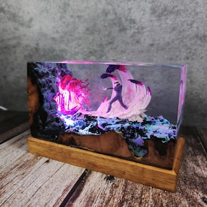 May include: A rectangular resin art piece featuring a miniature figure in a dynamic pose, set against a backdrop of illuminated, abstract forms in pink, purple, and blue. The artwork is mounted on a wooden base.