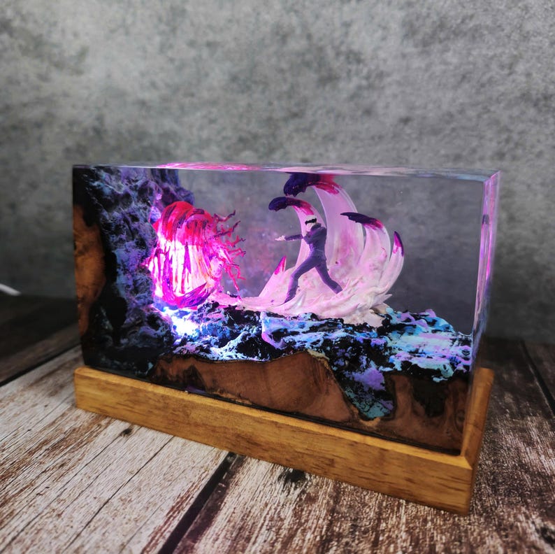 May include: A rectangular resin art piece featuring a miniature figure in a dynamic pose, set against a backdrop of illuminated, abstract forms in pink, purple, and blue. The artwork is mounted on a wooden base.