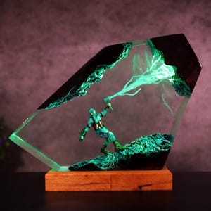 K&ouml;nnte beinhalten: Dekorative Resin-Skulptur mit einer Captain America-Figur mit Hammer, eingebettet in einen klaren, abgewinkelten Block. Die Skulptur hat einen Holzsockel und leuchtend gr&uuml;ne Akzente, die eine dynamische und beleuchtete Darstellung erzeugen.