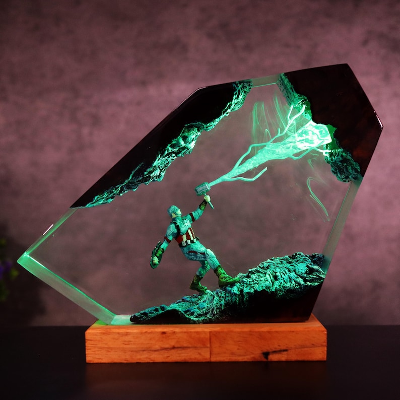 K&ouml;nnte beinhalten: Dekorative Resin-Skulptur mit einer Captain America-Figur mit Hammer, eingebettet in einen klaren, abgewinkelten Block. Die Skulptur hat einen Holzsockel und leuchtend gr&uuml;ne Akzente, die eine dynamische und beleuchtete Darstellung erzeugen.
