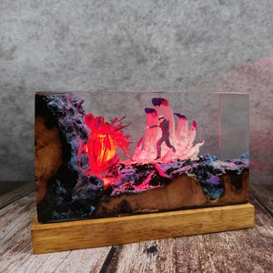 May include: A rectangular resin art piece with a wooden base. The artwork features a miniature figure running towards a glowing red and pink landscape. The resin is clear, showcasing the detailed scene with vibrant colors and textures.