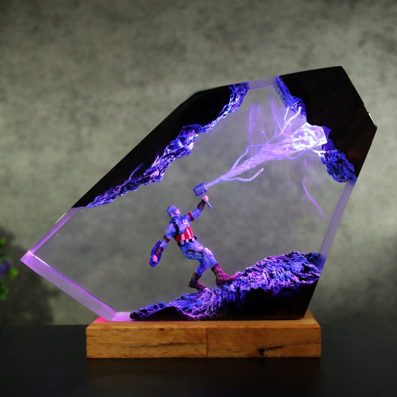 K&ouml;nnte beinhalten: Eine Captain America-Figur in einem transparenten, abgewinkelten Harzblock mit Holzsockel. Die Figur h&auml;lt einen Hammer, mit lila Blitzeffekten. Das Harz hat eine dunkle, strukturierte Oberseite und wird von violettem Licht beleuchtet.