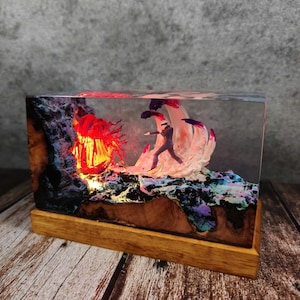 May include: A rectangular resin art piece featuring a miniature figure facing a fiery, red-lit structure. The base is wood, and the scene is encased in clear resin. The artwork has a fantasy theme with vibrant colors.