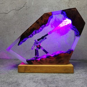 May include: A unique resin lamp featuring a robot figure with a weapon, set within a wooden and resin frame. The lamp emits a vibrant purple glow, with the robot figure standing on a textured base. The base is made of wood.