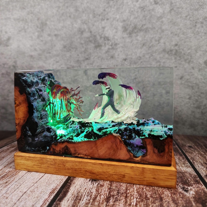 May include: A rectangular resin art piece featuring a miniature figure in a wave scene. The base is wood, and the resin has a layered effect with green and purple lighting. The figure is in a dark suit, and the waves are white and purple.
