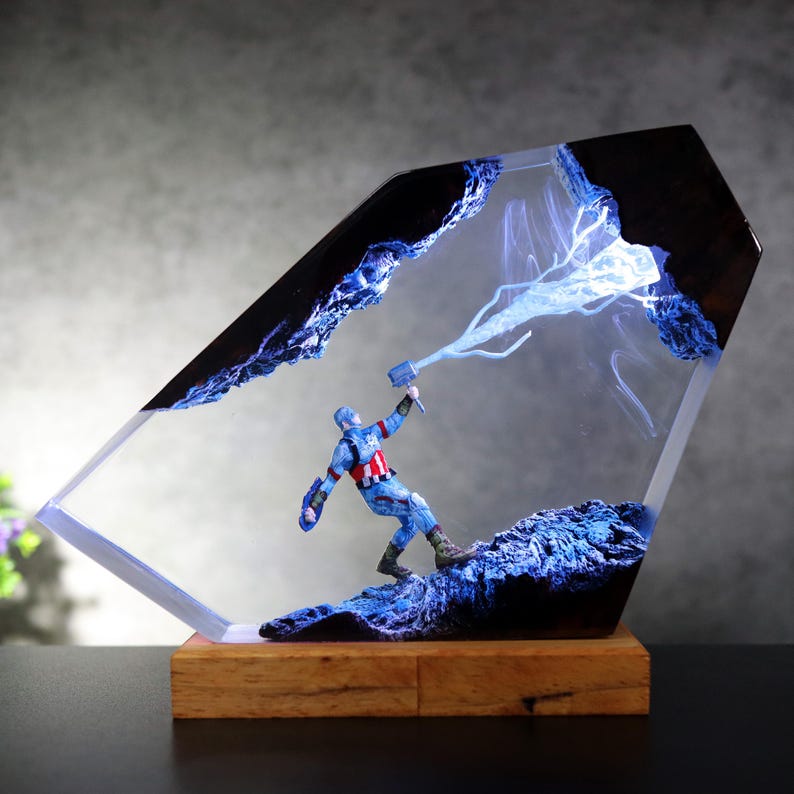 K&ouml;nnte beinhalten: Eine transparente Resin-Skulptur mit Captain America und seinem Hammer, vor dem Hintergrund blauer Blitze und dunkler Felsformationen. Die Skulptur ist auf einem Holzsockel montiert und erzeugt eine dynamische und detaillierte Darstellung.