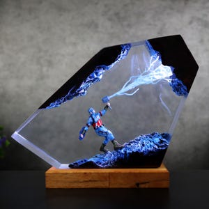 K&ouml;nnte beinhalten: Dekorative Resin-Skulptur mit Captain America und seinem Hammer in einem transparenten, abgewinkelten Rahmen. Das Design umfasst blaue Blitzeffekte und steht auf einem Holzsockel. Ein einzigartiges Kunstwerk.
