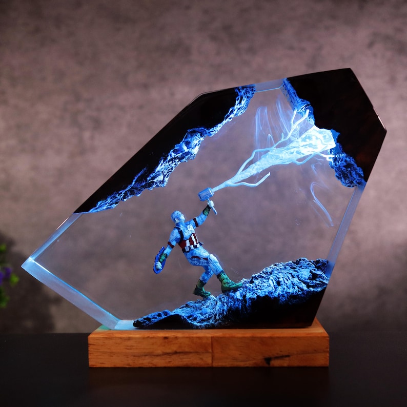 K&ouml;nnte beinhalten: Dekorative Resin-Skulptur mit einer Superheldenfigur, die einen Hammer h&auml;lt, umgeben von blauem Blitz und einer felsigen Landschaft. Die Skulptur ist in einem klaren, abgewinkelten Resinblock auf einem Holzsockel montiert.