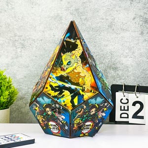 Könnte beinhalten: Eine facettenreiche, geometrische Lampe mit farbenfrohen Illustrationen von Anime-Charakteren. Die Lampe hat eine Diamantenform mit detaillierten Kunstwerken auf jeder Platte. Eine Fernbedienung und ein Kalender mit dem 25. Dezember sind sichtbar.