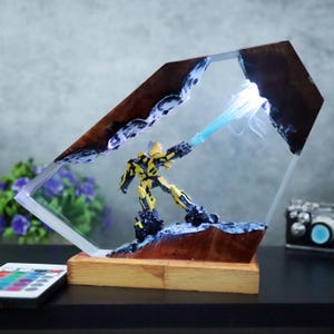 Könnte beinhalten: Eine gelb-schwarze Actionfigur ist in einem transparenten Harzblock mit Holzsockel eingeschlossen. Die Figur ist so positioniert, als würde sie einen blauen Energiestrahl abfeuern. Der Harzblock hat ein braun-weißes strukturiertes Design, und die Basis ist rechteckig.