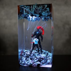 Könnte beinhalten: Ein rechteckiges Kunstwerk aus Kunstharz mit zwei Figuren in einer Kampfszene. Eine Figur trägt einen schwarzen Anzug mit einer roten Sense, die andere ein blaues Hemd. Das Harz hat blaue und weiße Akzente mit einem dunklen Holzrahmen.