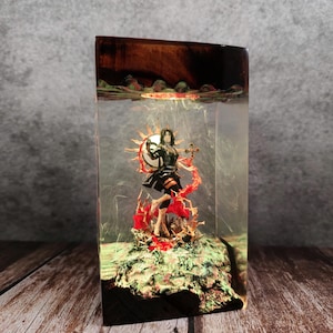 May include: A resin sculpture featuring a female figure with a shield and cross, set within a clear rectangular block. The figure is surrounded by red and orange accents, with a wooden top and base. The sculpture is approximately 4 inches tall.