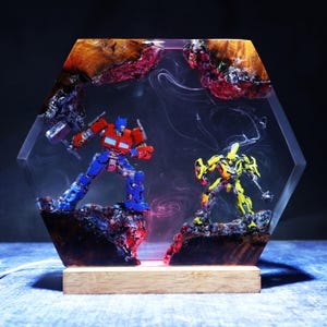 Może przedstawiać: Sześciokątna lampa z żywicy z figurkami Optimusa Prime'a i Bumblebee zatopionymi w przezroczystej żywicy i naturalnym drewnie. Scena zawiera wirujące efekty białego dymu, czerwone oświetlenie oraz jasną drewnianą podstawę.