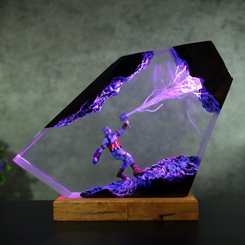 K&ouml;nnte beinhalten: Eine Captain America-Figur in einem transparenten, abgewinkelten Harzblock mit violetten Lichteffekten. Der Sockel ist ein Holzblock. Das Harz hat schwarze Akzente und einen violetten Blitzeffekt. Die Figur befindet sich in einer dynamischen Pose.
