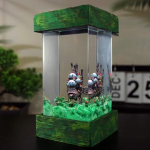 May include: A decorative terrarium with a miniature castle scene. The clear glass enclosure has a green, textured wooden base and top. Inside, a detailed model of a castle with blue and white accents sits on a green landscape. The terrarium is approximately 15 cm tall.