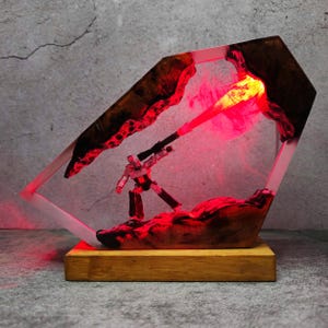 May include: A unique resin art piece featuring a small robot figure with a red laser beam. The artwork is encased in a clear, geometric frame with a wooden base. The interior is illuminated with red light.