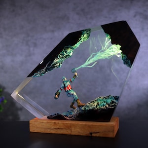 K&ouml;nnte beinhalten: Eine Captain America-Figur in einem transparenten Harzblock mit Holzsockel. Die Figur h&auml;lt einen Hammer und steht auf einer felsigen Oberfl&auml;che. Der Harzblock hat eine dunkle, strukturierte Oberseite und einen gr&uuml;nen Leuchteffekt.