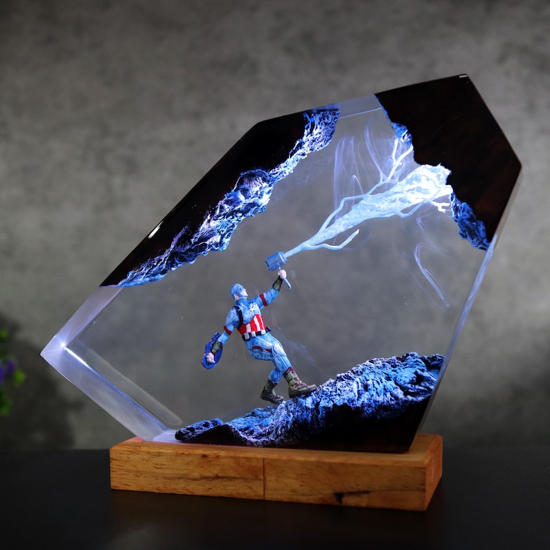 K&ouml;nnte beinhalten: Captain America-Figur in einem transparenten Harzblock mit Holzsockel. Das Harz hat blaue Akzente und einen Blitz-Effekt. Die Figur h&auml;lt einen Hammer und einen Schild.