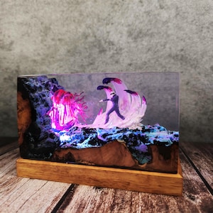 May include: A rectangular resin art piece featuring a miniature figure facing a glowing, abstract creature and wave-like forms. The artwork is set on a wooden base, with the scene illuminated by purple and blue lights, creating a fantasy theme.