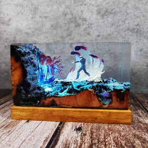 May include: A rectangular resin art piece featuring a miniature figure in a suit, set against a backdrop of stylized waves and a waterfall. The piece is encased in clear resin, with a wooden base. The color palette includes blues, purples, and browns.