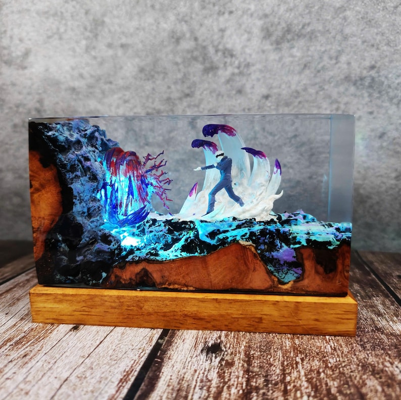 May include: A rectangular resin art piece featuring a miniature figure in a suit, set against a backdrop of stylized waves and a waterfall. The piece is encased in clear resin, with a wooden base. The color palette includes blues, purples, and browns.