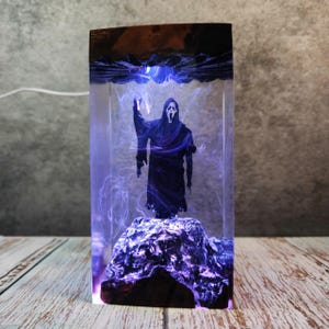 Puede incluir: Una pieza de arte de resina rectangular con una figura negra con una máscara de Scream, de pie sobre una base oscura y texturizada. La figura está encerrada en resina transparente con detalles morados y azules arremolinados.