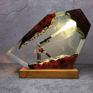 May include: A unique lamp featuring a robot figure encased in a clear resin structure. The resin is accented with red and brown tones, and the lamp sits on a wooden base. The robot is holding a weapon, and the lamp emits a bright light.