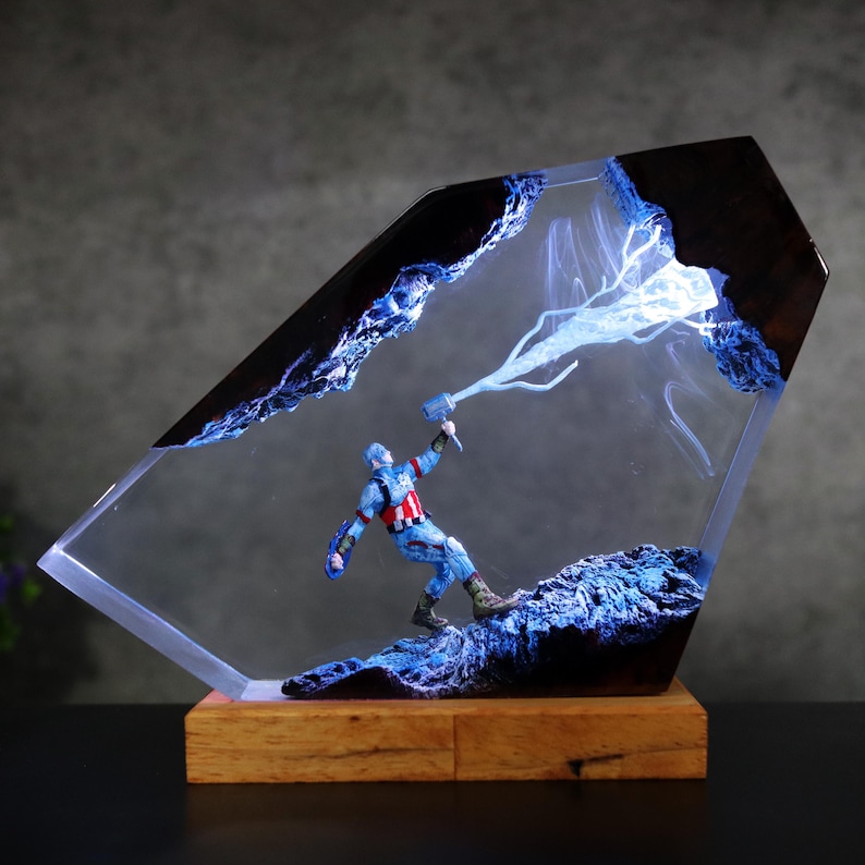 K&ouml;nnte beinhalten: Eine Captain America-Figur in einem transparenten Harzblock mit Holzsockel. Die Figur h&auml;lt einen Hammer und ist von blauem Blitz und einer blauen Felslandschaft umgeben. Der Harzblock hat einen dunklen, strukturierten Rand.