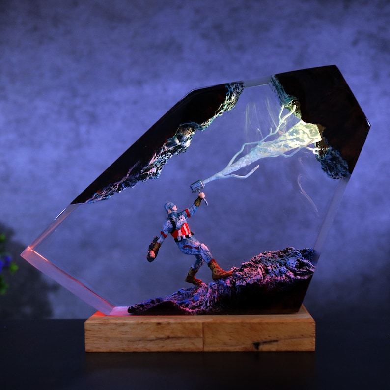 K&ouml;nnte beinhalten: Eine Captain America-Figur in einem transparenten, abgewinkelten Harzblock mit Holzsockel. Die Figur steht auf einer felsigen Oberfl&auml;che, mit einem Blitzeffekt dar&uuml;ber. Der Harzblock hat eine dunkle, strukturierte Oberseite.