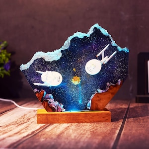 Puede incluir: Lámpara decorativa que representa una escena espacial con dos naves espaciales blancas y un sol amarillo sobre un fondo de galaxia azul y púrpura. La lámpara está colocada sobre una base de madera y tiene una luz azul que emana de la parte inferior.