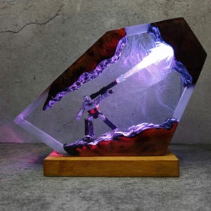 May include: A unique, illuminated resin sculpture featuring a robot figure in a dynamic pose. The sculpture is encased in a clear, geometric form with wood and purple accents. The base is made of wood, and the interior is lit with a bright light.