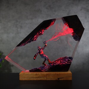 K&ouml;nnte beinhalten: Eine Captain America-Figur in einem transparenten, abgewinkelten Harzblock mit roter Innenbeleuchtung. Die Figur ist mit einem Hammer posiert und steht auf einem Holzsockel. Das Harz hat schwarze und rote Akzente.