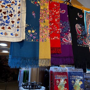 Peut inclure: Étalage de foulards colorés ornés de motifs floraux et cachemire complexes. Les foulards, de couleurs variées comme le bleu, le jaune, le rouge, le violet et le noir, sont suspendus à une barre métallique. Certains ont des franges.