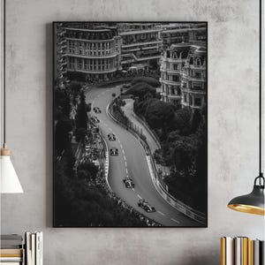 May include: Black and white framed print of a Formula 1 race. The image captures race cars speeding along a winding track through a city, with buildings and spectators visible. The artwork is a high-angle shot.