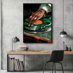 Pode incluir: Uma impress&atilde;o art&iacute;stica emoldurada retrata a m&atilde;o de um DJ com an&eacute;is e pulseiras douradas em um disco de vinil verde. A obra de arte &eacute; colocada contra uma parede de concreto cinza, com uma mesa, uma l&acirc;mpada e uma cadeira em primeiro plano.