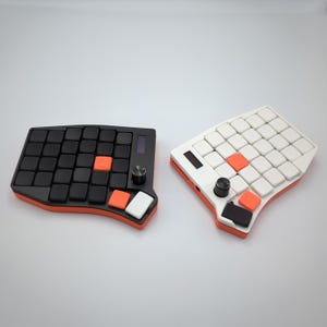 Könnte beinhalten: Zwei ergonomische mechanische Tastaturen. Eine ist schwarz mit orangefarbenen Akzenten und die andere ist weiß mit orangefarbenen Akzenten. Jede hat ein Raster aus quadratischen Tasten, eine zentrale orangefarbene Taste, ein Rad und ein kleines Display.