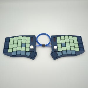 May include: A split ergonomic keyboard with a dark blue base, featuring light green and blue keycaps. The keyboard is connected by a blue cable and has silver knobs and small screens.