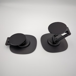 May include: Two matte black adhesive phone mounts with circular magnetic heads and square base plates. One mount is folded flat, while the other is extended with an adjustable hinged arm, both featuring a smooth, solid black finish throughout.