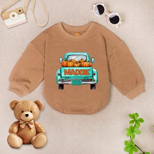 May include: A tan-colored baby onesie with a crew neck and long sleeves. The front features a teal truck graphic filled with orange pumpkins, the name "MADDIE", and the number "2025". A teddy bear, sunglasses, and a toy camera are also visible.