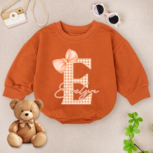 May include: An orange baby onesie with long sleeves and a round neckline. The onesie features a large letter "E" in a gingham pattern, a bow, and the name "Evelyn" in script. A teddy bear, sunglasses, and a toy camera are also in the image.