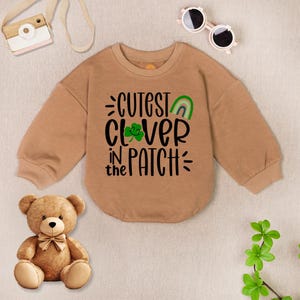 May include: A tan-colored baby bodysuit with long sleeves. The front of the bodysuit features the text "Cutest Clover in the Patch" in black, with a green shamrock and rainbow graphic. A teddy bear and sunglasses are in the background.