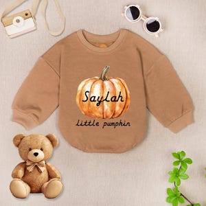 May include: A tan-colored baby bodysuit with a watercolor pumpkin design. The pumpkin has the name "Saylah" and the phrase "Little pumpkin" printed on it. The bodysuit is surrounded by a teddy bear, sunglasses, and a toy camera.