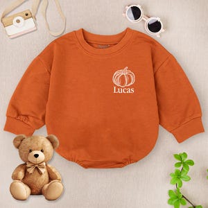 May include: An orange baby bodysuit with a white pumpkin graphic and the name "Lucas" printed below it. The long-sleeved outfit has ribbed cuffs and a round neckline. A teddy bear, toy camera, sunglasses, and greenery are also in the image.