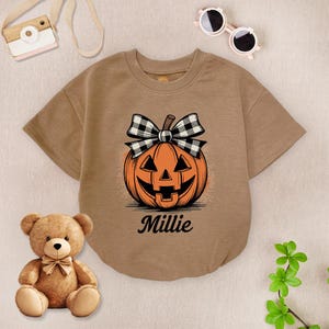May include: A tan short-sleeved baby bodysuit featuring a smiling jack-o'-lantern graphic with a black and white plaid bow. The name "Millie" is printed below the pumpkin. A teddy bear, toy camera, and sunglasses are also in the image.
