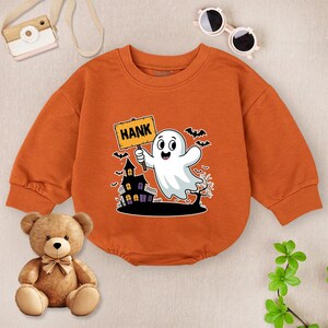 May include: Orange Halloween-themed baby bodysuit with a graphic of a friendly ghost holding a sign that says "HANK". The design includes a haunted house, bats, and a tree. The long-sleeved bodysuit is made of soft material.