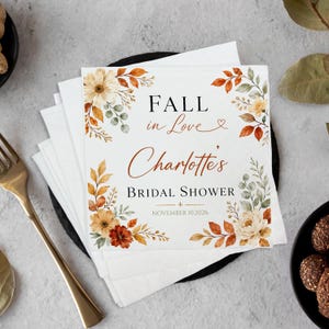 May include: A square white paper napkin featuring a floral border with autumn-colored leaves and flowers in shades of orange, brown, and green. The text reads FALL in Love, Charlotte's BRIDAL SHOWER, and the date NOVEMBER 10, 2026.