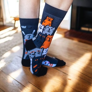 Pode incluir: Um par de meias cinza escuro com um padrão repetido de gatos de desenho animado em laranja, cinza e preto. As meias têm calcanhares e dedos pretos. Os gatos têm a letra "T" no peito.