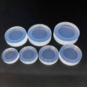 Puede incluir: Colección de siete moldes circulares de silicona transparente de varios tamaños. Cada molde tiene un borde exterior elevado y un centro hundido. Los moldes están dispuestos sobre una superficie oscura, mostrando su transparencia.