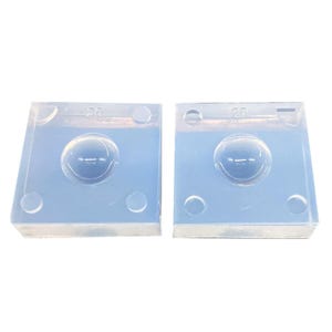 Puede incluir: Dos moldes cuadrados de silicona transparente con una hendidura redondeada en forma de burbuja en el centro. Cada molde tiene el número "20" grabado en la parte superior. Los moldes están diseñados para la elaboración o creación de objetos esféricos.